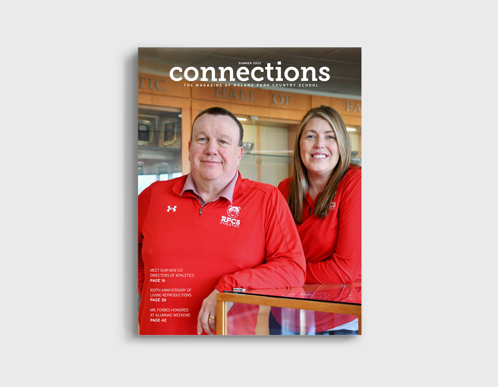 Cover of Connections Alumni Magazine featuring a middle-aged man and woman wearing red Roland Park Country School athletic shirts standing in front of a trophy case that says "Hall of Fame" above it.